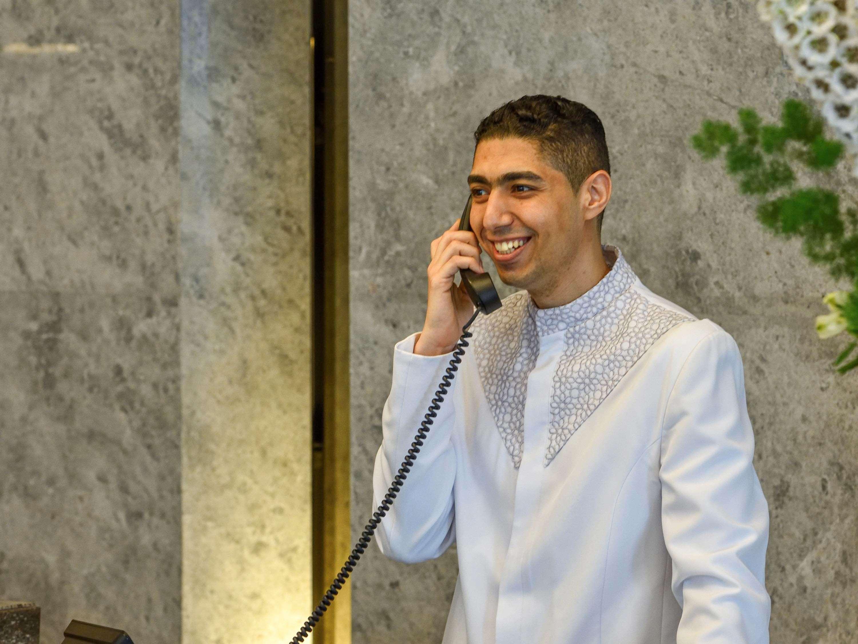 Grand Mercure Dubai City Exterior photo The photo shows a young man dressed in a formal white outfit with decorative details around the collar. He is smiling and is speaking on a vintage-style telephone. The background appears to have a marble texture, and there are some plants or floral a