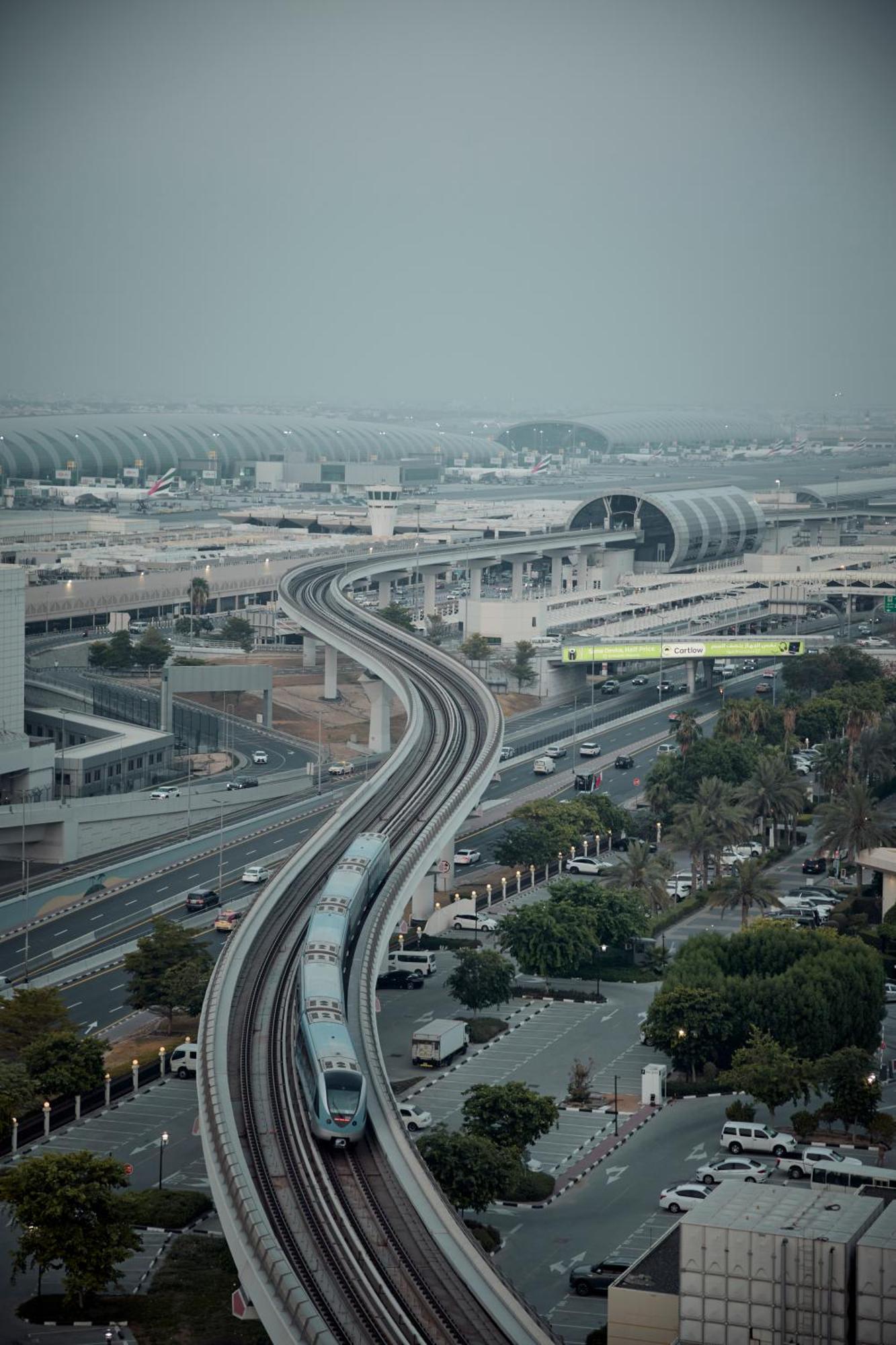 Grand Mercure Dubai City Exterior photo The photo depicts an urban landscape featuring a monorail or light rail system curving along a track above ground. Below the elevated rail, there are roads with vehicles and greenery along the sides. The background shows modern architecture, likely r