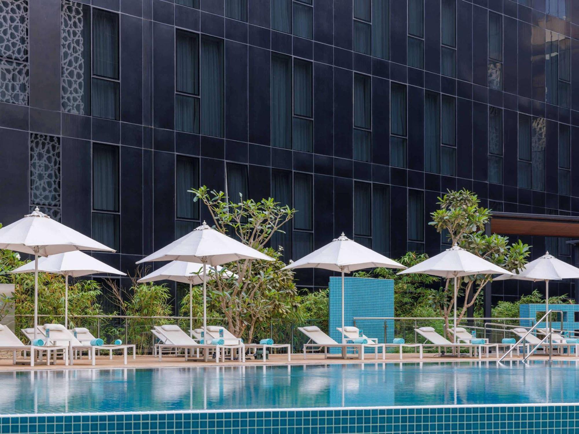Grand Mercure Dubai City Exterior photo The photo depicts a modern swimming pool area surrounded by lounge chairs and umbrellas. The pool has a turquoise color and is situated in front of a sleek, dark building with large windows. Several white umbrellas provide shade for the lounge chairs